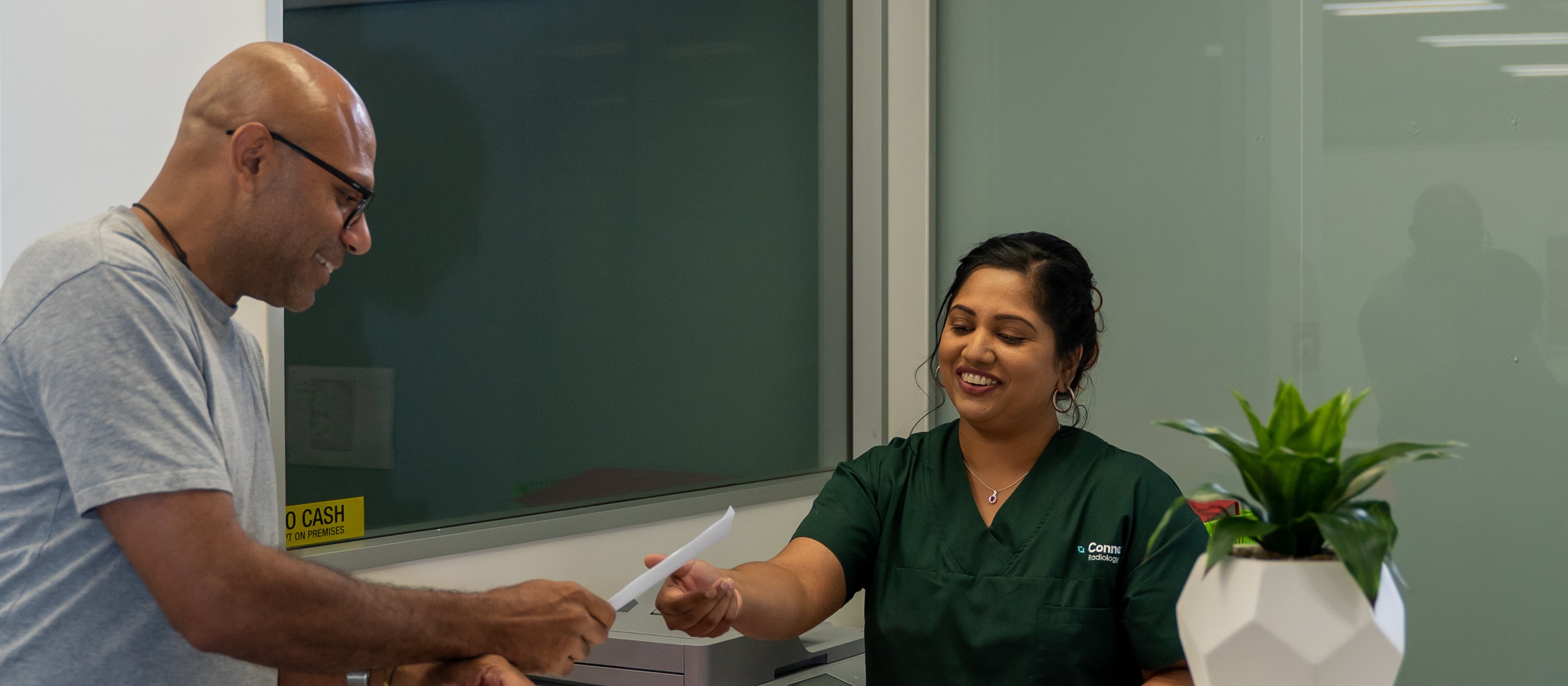 Image shows a man at the reception desk of Connect Radiology, with woman behind the counter reaching for his referral papers