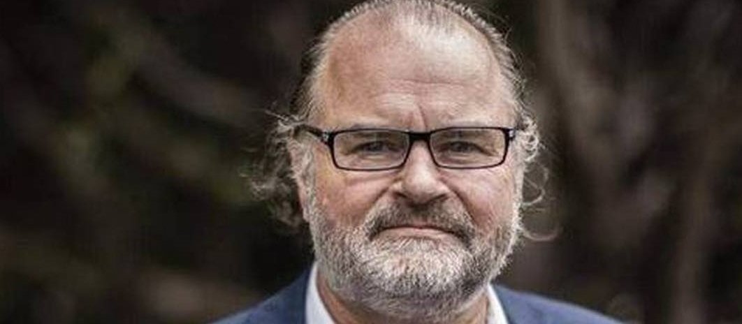 Image shows man in pale shirt and blue blazer, with glasses and a hei tiki bone necklace, and short greay beard, looking at the camera