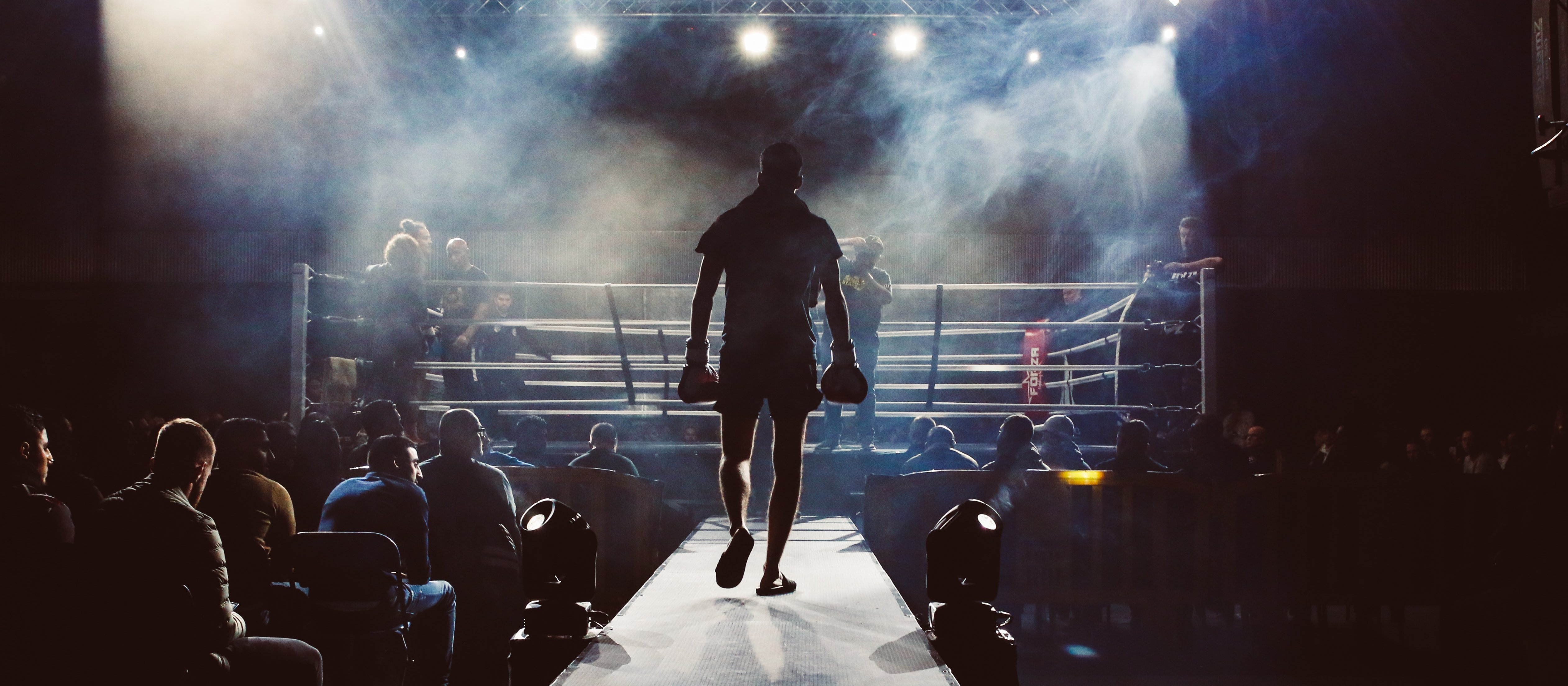 Image shows man walking towards a boxing ring, ready for a match, with lights and smoke in the background