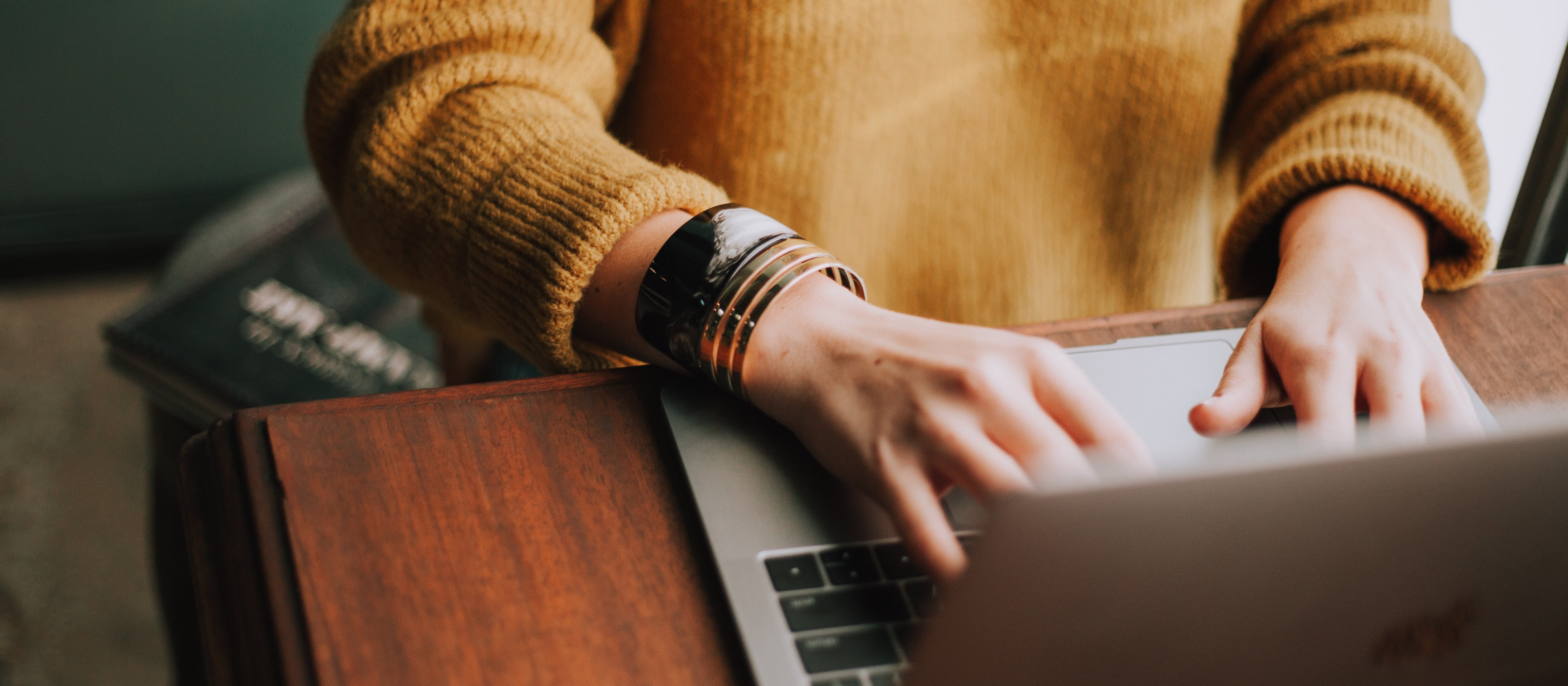 A person in a mustard-coloured jumper and chunky bracelets typing on a laptop at a dark wood desk