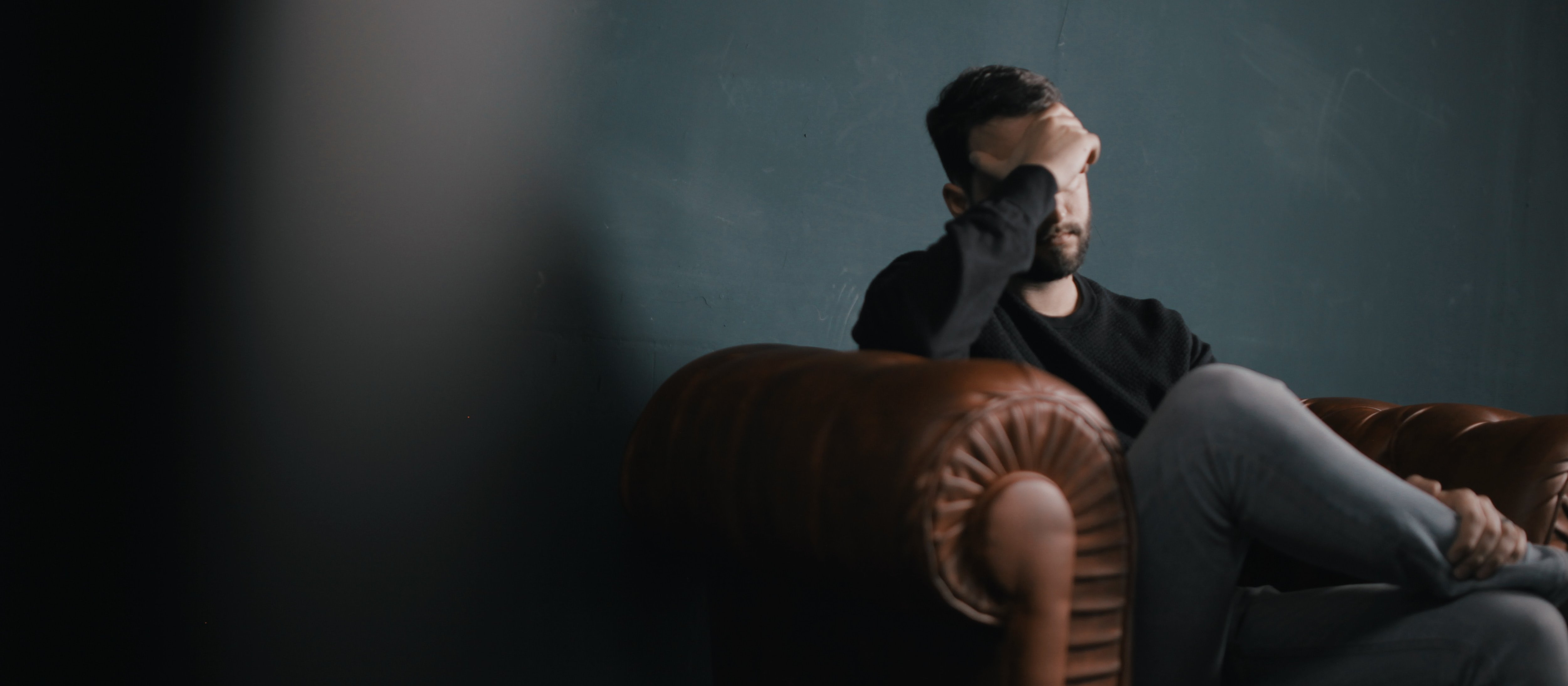 Image shows man sitting on leather couch, with his hand to his forehead