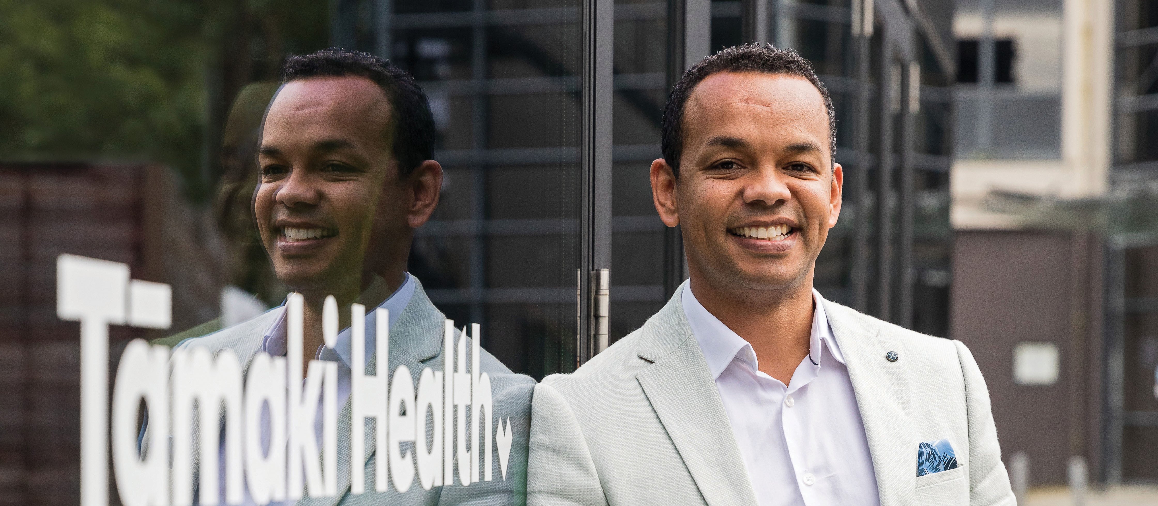 Image shows man in light blazer and white shirt leaning against a reflective window with the sign Tāmaki Health in white writing