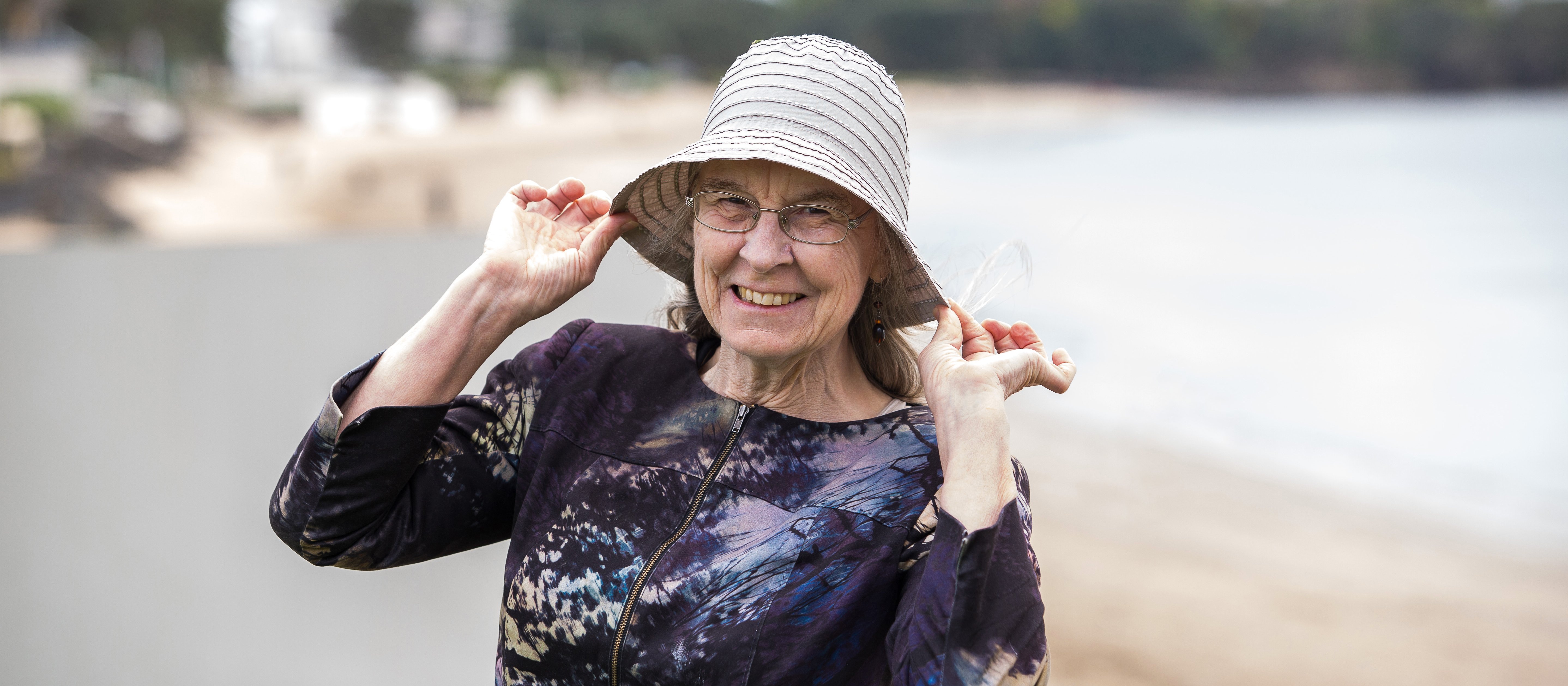 Amanda Oakley holds the edges of her cream hat, smiling at the camera, beach in background