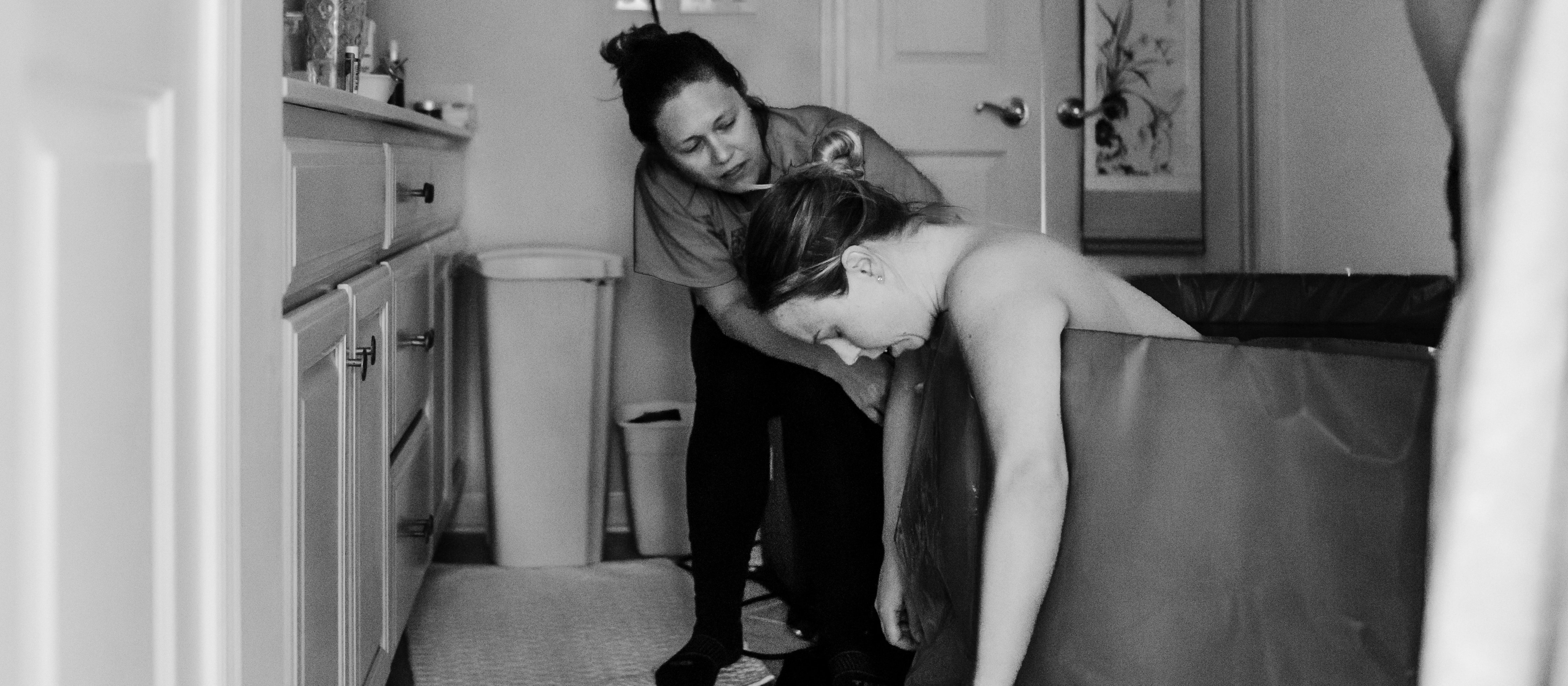 Black and white photo shows a woman, arms hanging out of a birthing pool, in a bathroom, supported by her midwife