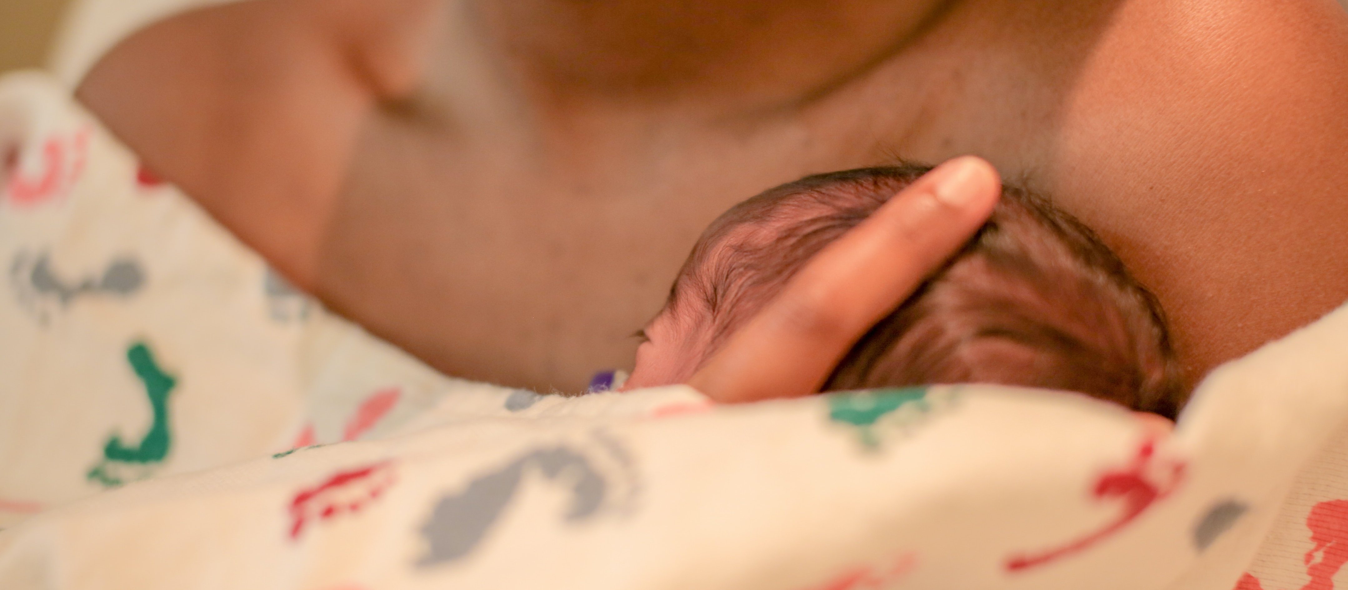 Image shows the head of a newborn baby poking out of a blanket wrapped around the chest of their mother, whose shoulders and chin are shown