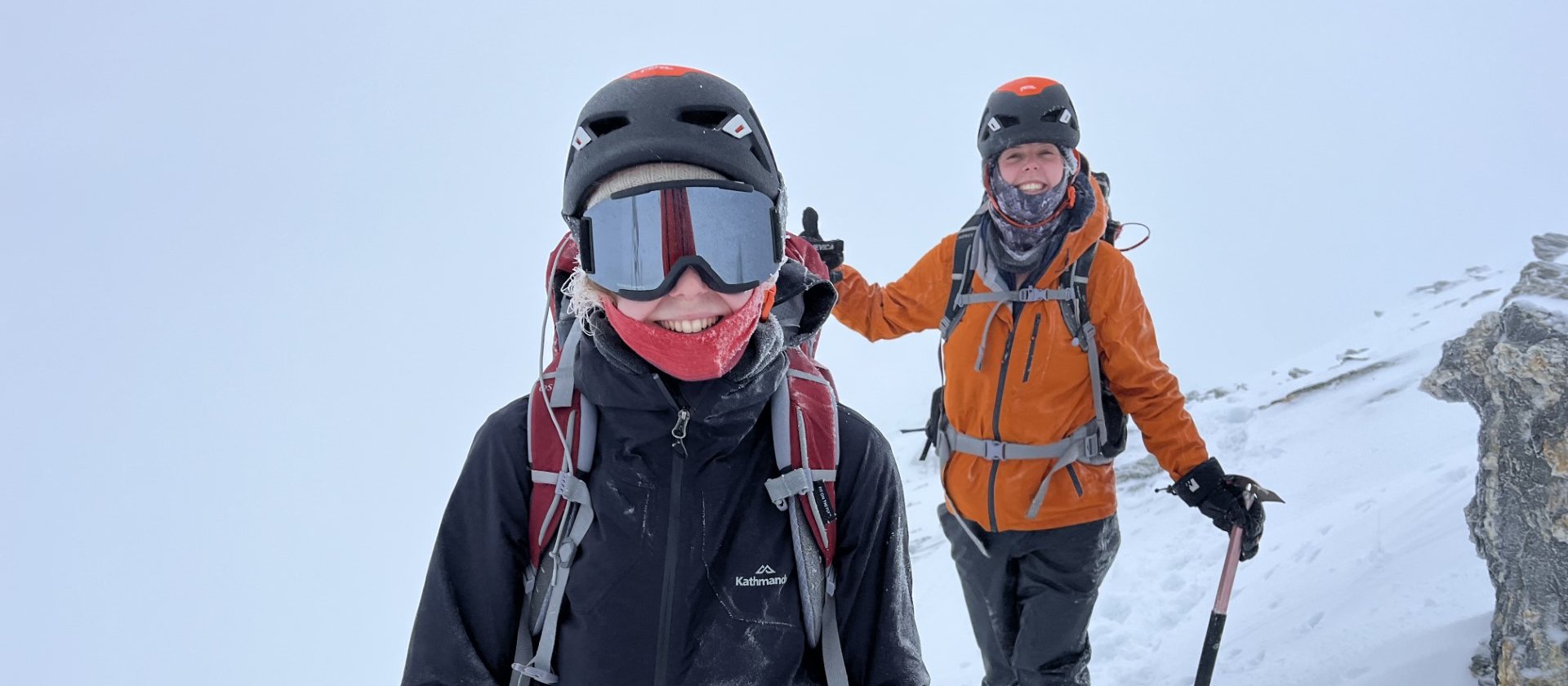 Two women in mountaineering gear hiking on a snowy mountain, with snow falling around them