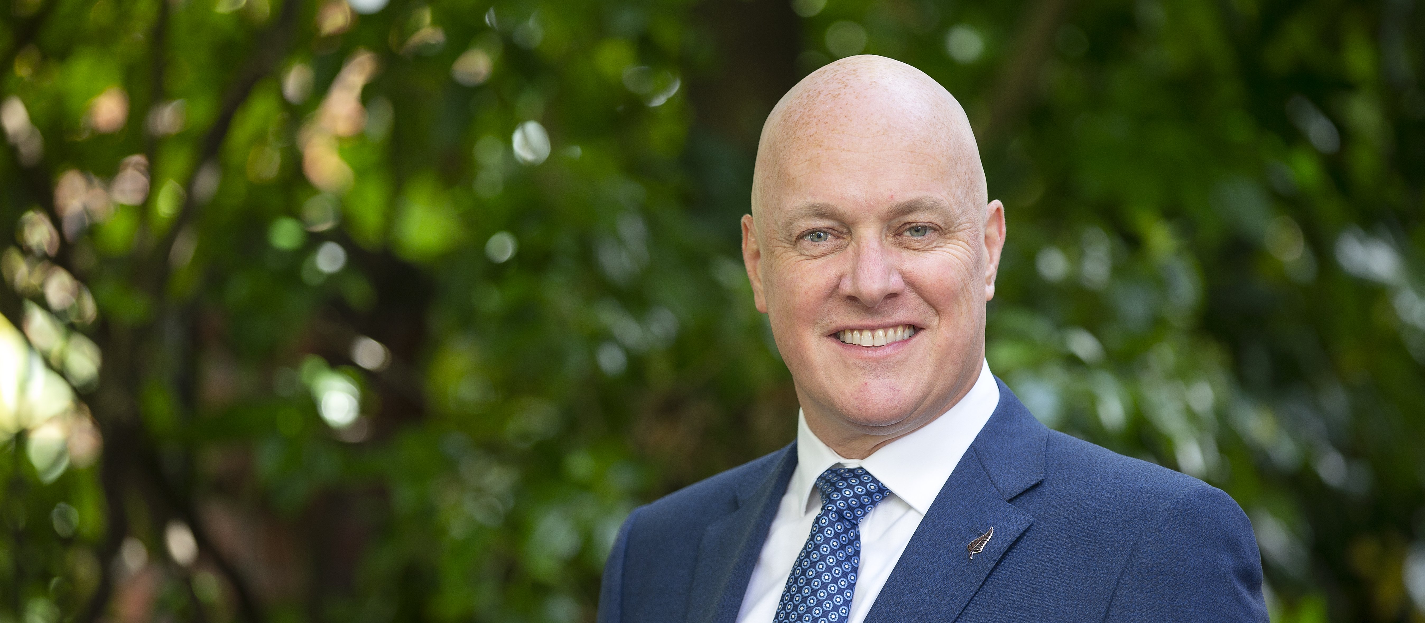 Christopher Luxon, in dark blue suit, white shirt and blue tie, trees in background