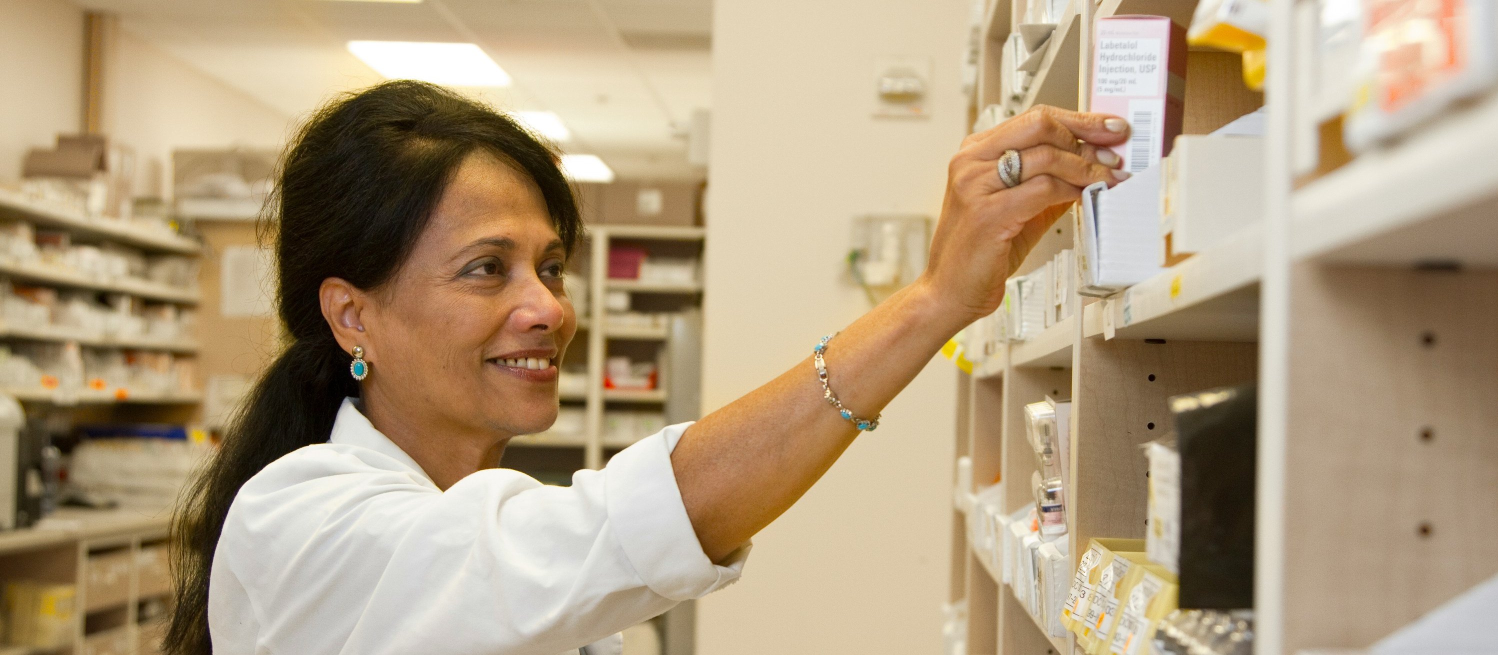 Woman with long dark hair lifting a medicine box from a shelf. Background shows shelves full of medicines at a pharmacy