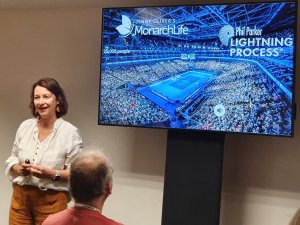 Image shows a woman, Jenny Oliver, standing beside a television screen during a presentation on the Lightning Process