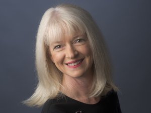 Woman with blonde hair and black top, standing sideways and looking towards the camera, smiling