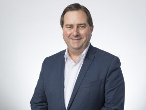 Image shows a man in a white shirt and navy blazer, short dark hair, smiling at the camera