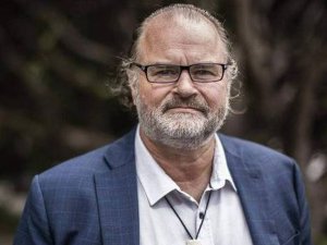 Image shows man in pale shirt and blue blazer, with glasses and a hei tiki bone necklace, and short greay beard, looking at the camera