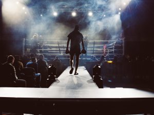 Image shows man walking towards a boxing ring, ready for a match, with lights and smoke in the background