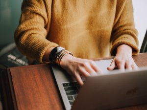 A person in a mustard-coloured jumper and chunky bracelets typing on a laptop at a dark wood desk