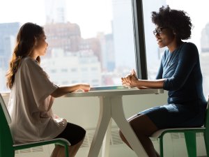 Two women sitting across from each other at a round white table in front of a window, both have hands on the table