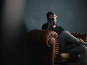 Image shows man sitting on leather couch, with his hand to his forehead