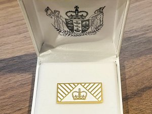 A gold lapel pin, with a crown in the centre, sits on a white display case with the NZ coat of arms on the underside of the lid