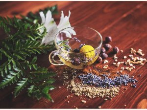 Image shows a clear glass cup of herbal tea, with flowers inside, on a wooden table strewn with herbs, leaves, and seeds