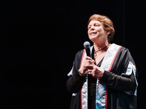 Image shows woman with short hair holding a microphone while giving a speech, wearing a ceremonial gown, on a black background