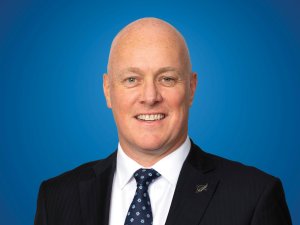 Image shows bald man in black suit, white shirt and blue tie, smiling at camera, against a blue background