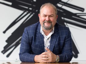 Man with beard sits at a table, hands clasped in front of him, wearing a blue blazer. A black Spark logo in the background