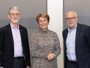 Image shows a woman in printed dress standing between two men in suits