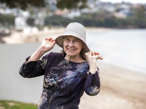 Amanda Oakley holds the edges of her cream hat, smiling at the camera, beach in background