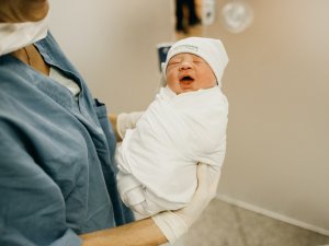 A newborn baby wrapped in a white swaddle and hat, being held by an adult in blue shirt, hospital room