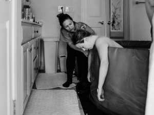 Black and white photo shows a woman, arms hanging out of a birthing pool, in a bathroom, supported by her midwife