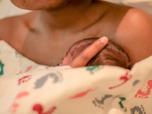 Image shows the head of a newborn baby poking out of a blanket wrapped around the chest of their mother, whose shoulders and chin are shown