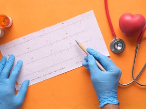 Image shows gloved hands checking an ECG report, with stethoscope and heart image on an orange background