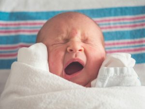 A newborn yawns, they're swaddled in white and lying on a white blanket with blue and pink stripes