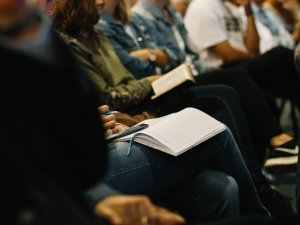 Image shows a row of seated people books and notepads on their knees, taking notes at a conference