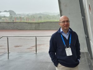 Image shows bald man in navy jumper standing outside conference centre, grey rainy background