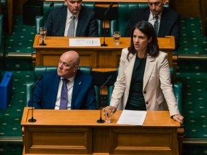 Minister Erica Stanford stands while speaking in Parliament, Prime Minister Christopher Luxon sits beside her